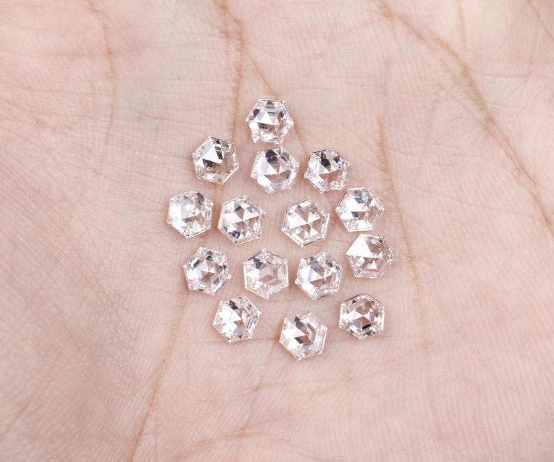Puede incluir: Una colecci&oacute;n de catorce peque&ntilde;as piedras preciosas transparentes de corte hexagonal con superficies facetadas. Cada piedra presenta una forma geom&eacute;trica de seis lados con una parte superior puntiaguda, reflejando la luz a trav&eacute;s de sus m&uacute;ltiples facetas pulidas, dispuestas en un grupo suelto.