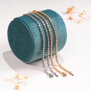 K&ouml;nnte beinhalten: Drei zarte Tennisarmb&auml;nder in Silber, Gold und Ros&eacute;gold, jedes mit funkelnden Edelsteinen besetzt. Die Armb&auml;nder sind &uuml;ber einem t&uuml;rkisfarbenen Samt-Schmuckst&auml;nder drapiert, mit Trockenblumen im Hintergrund.