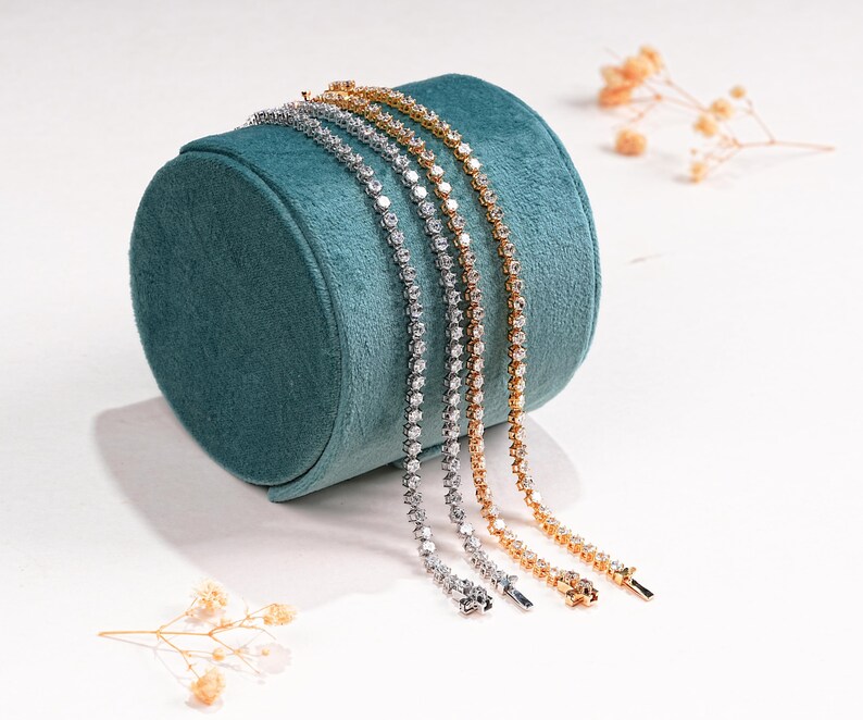 K&ouml;nnte beinhalten: Drei zarte Tennisarmb&auml;nder in Silber, Gold und Ros&eacute;gold, jedes mit funkelnden Edelsteinen besetzt. Die Armb&auml;nder sind &uuml;ber einem t&uuml;rkisfarbenen Samt-Schmuckst&auml;nder drapiert, mit Trockenblumen im Hintergrund.
