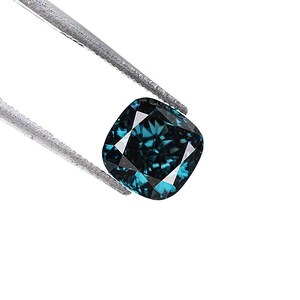 May include: A cushion-cut, teal-colored gemstone is held by a pair of silver tweezers against a white background. The gemstone has a square shape with rounded corners and multiple facets that reflect light.