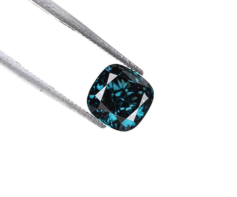 May include: A cushion-cut, teal-colored gemstone is held by a pair of silver tweezers against a white background. The gemstone has a square shape with rounded corners and multiple facets that reflect light.