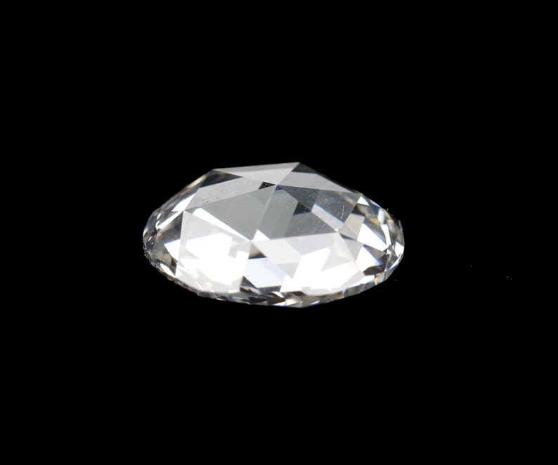 May include: A clear, faceted gemstone with a brilliant cut, resting against a black background. The gem's multiple facets reflect light, creating a sparkling effect. The overall shape is oval.