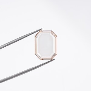 May include: An emerald-cut gemstone, held by tweezers, against a white background. The gem has a pale peach hue with a rectangular shape and a series of facets. The tweezers are silver-colored.