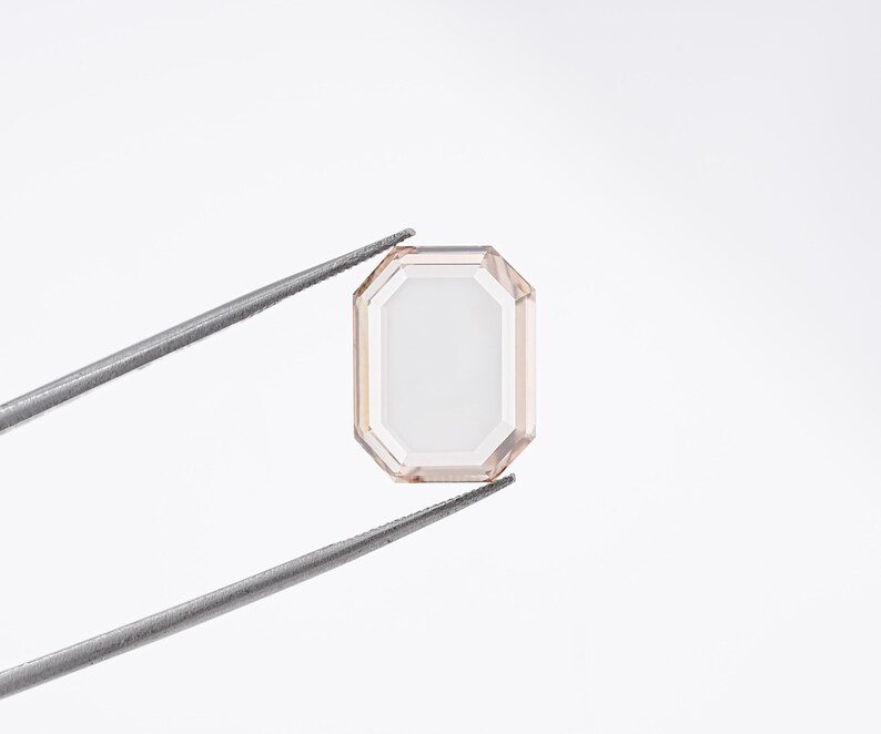 May include: An emerald-cut gemstone, held by tweezers, against a white background. The gem has a pale peach hue with a rectangular shape and a series of facets. The tweezers are silver-colored.