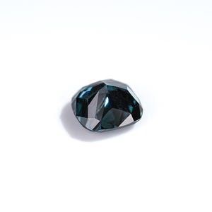 May include: A faceted, teal-colored gemstone with a unique, irregular shape. The gem reflects light, showcasing its multiple facets and internal depth. The stone is set against a plain white background, highlighting its color and form.