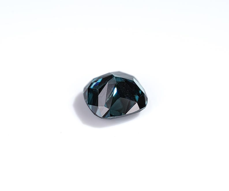 May include: A faceted, teal-colored gemstone with a unique, irregular shape. The gem reflects light, showcasing its multiple facets and internal depth. The stone is set against a plain white background, highlighting its color and form.