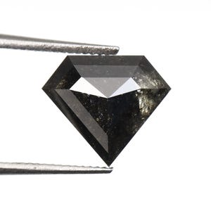 Peut inclure: Un diamant gris foncé facetté, tenu par une pince argentée. Le diamant a une forme géométrique avec de multiples facettes, reflétant la lumière. La gemme semble être une pierre naturelle non taillée, avec une texture mouchetée unique.