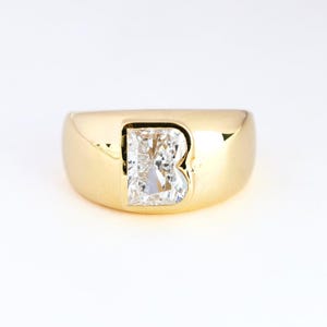 Könnte beinhalten: Ein goldener Ring mit einem großen, rechteckigen, klaren Edelstein in der Mitte. Der Ring hat ein breites Band und eine polierte Oberfläche, die das Licht reflektiert. Der Edelstein ist so geschliffen, dass er maximal glänzt.