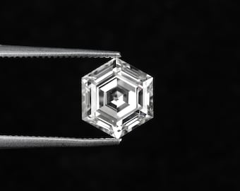 May include: A faceted, hexagonal-cut diamond held by tweezers against a black background. The diamond has a complex internal structure with multiple facets, reflecting light. The overall appearance is clear and brilliant.