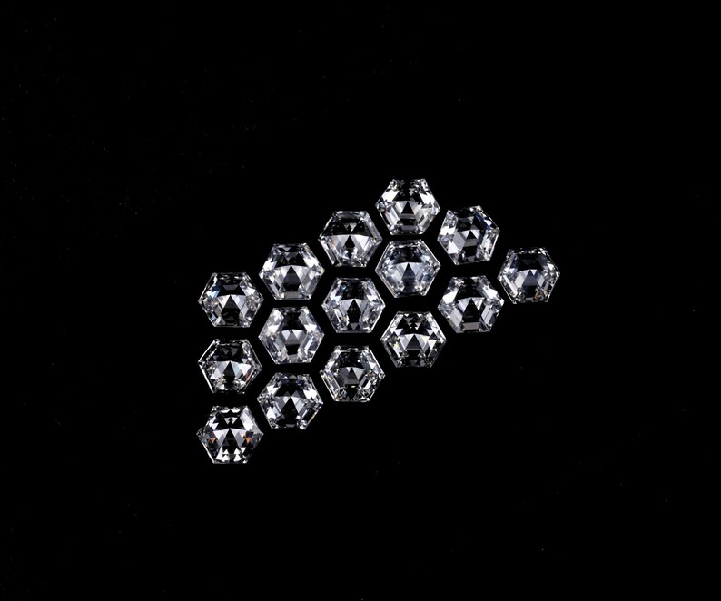 Puede incluir: Una colecci&oacute;n de catorce piedras preciosas sueltas, transparentes, incoloras y de corte hexagonal dispuestas en un grupo. Cada piedra presenta una superficie facetada que refleja la luz, creando un aspecto brillante y centelleante sobre un fondo negro.