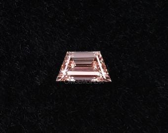 May include: A faceted, trapezoid-cut gemstone with a light peach hue. The gem features a series of parallel, horizontal facets, creating a reflective surface. The gemstone is set against a dark, solid background.
