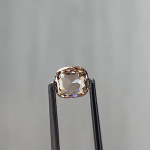 Może przedstawiać: Jasnobrązowy kamień szlachetny o szlifie poduszkowym jest trzymany przez czarną pęsetę. Kamień szlachetny ma wiele faset, które odbijają światło, tworząc efekt iskrzenia. Tło jest jednolite, neutralne szare.