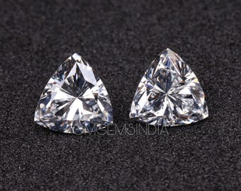 Può includere: Due gemme triangolari con taglio a brillante, con superfici sfaccettate. Le gemme chiare riflettono la luce, su uno sfondo scuro e strutturato. L'immagine ravvicinata evidenzia i tagli intricati e la chiarezza delle pietre.