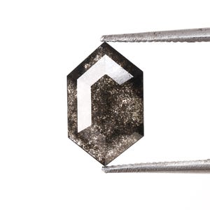 Może przedstawiać: Czarny diament o sześciokątnym kształcie z białym, geometrycznym wzorem w środku. Diament jest trzymany przez pęsetę.