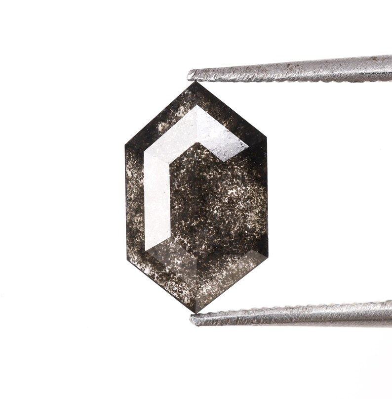 Może przedstawiać: Czarny diament o sześciokątnym kształcie z białym, geometrycznym wzorem w środku. Diament jest trzymany przez pęsetę.