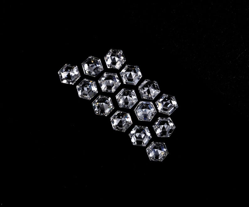 Puede incluir: Una colecci&oacute;n de catorce piedras preciosas transparentes e incoloras, con corte hexagonal y dispuestas en grupo. Cada piedra presenta una superficie facetada que refleja la luz, creando un aspecto brillante y centelleante sobre el fondo negro.