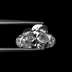 Könnte beinhalten: Ein klarer, wolkenförmiger Diamant, der von einer Pinzette vor einem schwarzen Hintergrund gehalten wird. Der Diamant ist facettiert und reflektiert das Licht. Die Pinzette ist dunkelgrau.
