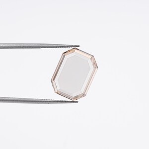 May include: A faceted, rectangular gemstone with a light peach hue, held by silver tweezers against a white background. The gem's edges are a darker peach, framing a lighter center. The overall shape is a modified octagon.