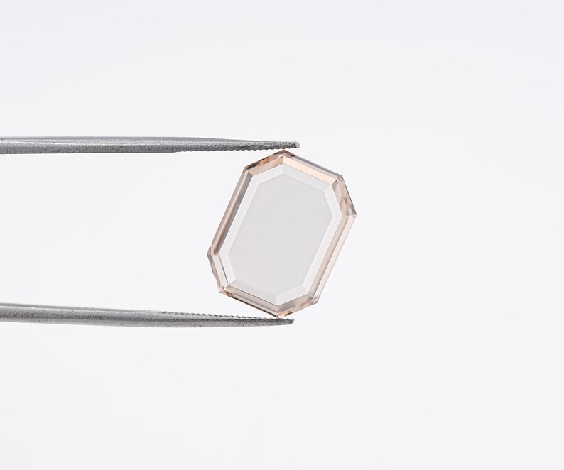 May include: A faceted, rectangular gemstone with a light peach hue, held by silver tweezers against a white background. The gem's edges are a darker peach, framing a lighter center. The overall shape is a modified octagon.