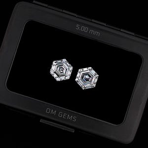 Puede incluir: Dos piedras preciosas transparentes de corte hexagonal exhibidas dentro de un estuche rectangular negro. El estuche presenta el texto OM GEMS y una medida de 5,00 mm impresa. Las piedras muestran un dise&ntilde;o geom&eacute;trico facetado.
