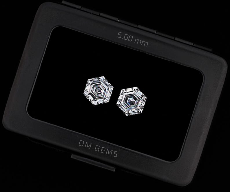Puede incluir: Dos piedras preciosas transparentes de corte hexagonal exhibidas dentro de un estuche rectangular negro. El estuche presenta el texto OM GEMS y una medida de 5,00 mm impresa. Las piedras muestran un dise&ntilde;o geom&eacute;trico facetado.