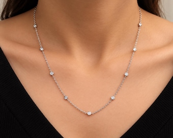 May include: A silver-toned chain necklace featuring multiple small, round, clear stones set in individual bezel settings spaced evenly along the length of the chain. The necklace rests against the neck, showcasing a delicate and refined design.