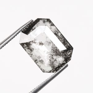 Könnte beinhalten: Ein facettierter, rechteckiger Edelstein mit Salz-und-Pfeffer-Aussehen, gehalten von einer Pinzette. Der Edelstein hat eine klare, reflektierende Oberfläche mit dunklen Einschlüssen, die ein einzigartiges Muster erzeugen. Der Hintergrund ist weiß.