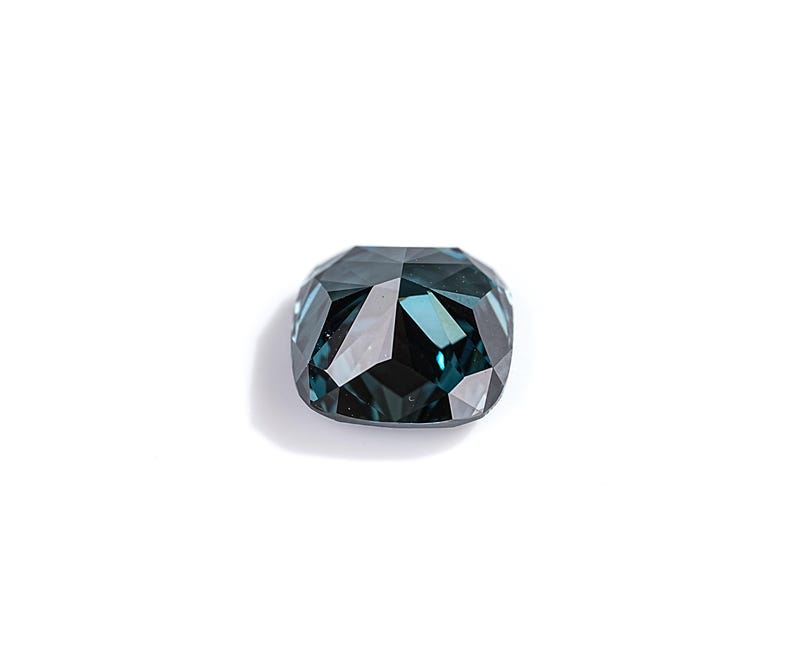 May include: A faceted, cushion-cut gemstone with a deep teal color. The gem is set against a white background, showcasing its multiple reflective surfaces. The overall shape is a rounded rectangle, with a high degree of clarity.
