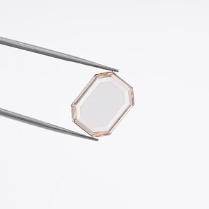May include: A faceted, light-colored gemstone with an octagonal shape is held by a pair of silver tweezers against a white background. The gem has a clear, reflective center and a subtle, warm-toned border.