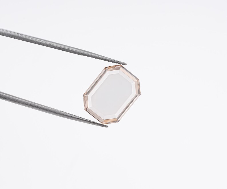 May include: A faceted, light-colored gemstone with an octagonal shape is held by a pair of silver tweezers against a white background. The gem has a clear, reflective center and a subtle, warm-toned border.