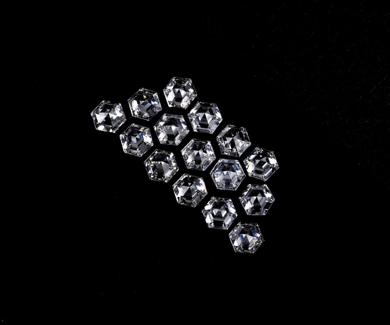 Puede incluir: Una colecci&oacute;n de catorce piedras preciosas transparentes e incoloras, con corte hexagonal y dispuestas en grupo. Cada piedra presenta una superficie facetada que refleja la luz, creando un aspecto brillante y centelleante sobre el fondo negro.