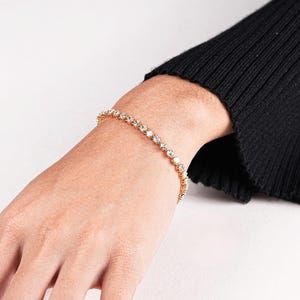 K&ouml;nnte beinhalten: Ein goldfarbenes Armband mit einer Reihe klarer, runder Edelsteine. Das Armband wird an einem hellh&auml;utigen Handgelenk getragen, im Hintergrund ein schwarzer &Auml;rmel. Die Edelsteine sind gleichm&auml;&szlig;ig verteilt und reflektieren das Licht.