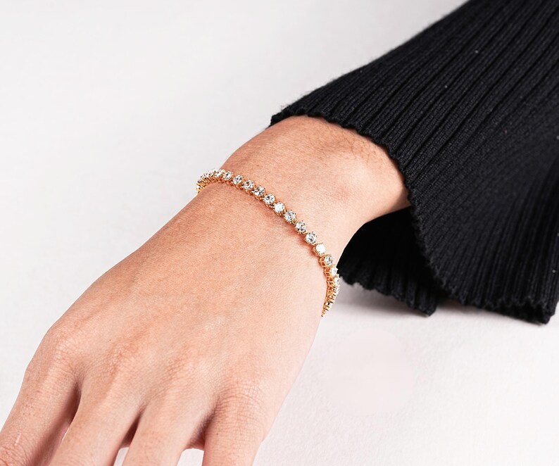 K&ouml;nnte beinhalten: Ein goldfarbenes Armband mit einer Reihe klarer, runder Edelsteine. Das Armband wird an einem hellh&auml;utigen Handgelenk getragen, im Hintergrund ein schwarzer &Auml;rmel. Die Edelsteine sind gleichm&auml;&szlig;ig verteilt und reflektieren das Licht.