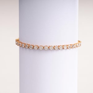 K&ouml;nnte beinhalten: Ein goldfarbenes Tennisarmband mit zahlreichen runden, klaren Edelsteinen. Das Armband wird auf einer wei&szlig;en zylindrischen Oberfl&auml;che pr&auml;sentiert, die sein zartes Design und seinen Glanz hervorhebt. Dieses Schmuckst&uuml;ck ist ein klassisches und elegantes Accessoire.