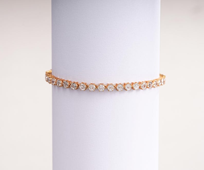 K&ouml;nnte beinhalten: Ein goldfarbenes Tennisarmband mit zahlreichen runden, klaren Edelsteinen. Das Armband wird auf einer wei&szlig;en zylindrischen Oberfl&auml;che pr&auml;sentiert, die sein zartes Design und seinen Glanz hervorhebt. Dieses Schmuckst&uuml;ck ist ein klassisches und elegantes Accessoire.