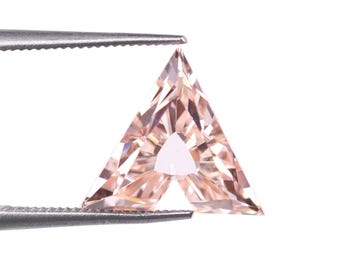 May include: A faceted, peach-colored gemstone held by silver tweezers. The gemstone is a unique triangular shape with a smaller triangle cut out of the center. The gem has many facets that reflect light.