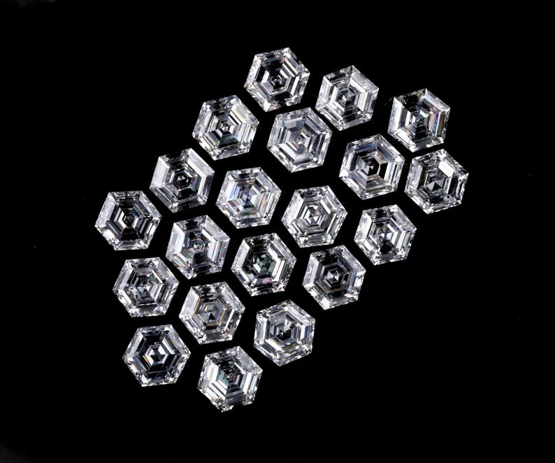 Puede incluir: Una colecci&oacute;n de diecinueve piedras preciosas transparentes e incoloras, con corte hexagonal y dispuestas en grupo. Cada piedra presenta una superficie facetada con un patr&oacute;n geom&eacute;trico que refleja la luz, creando un aspecto brillante y centelleante sobre un fondo negro.