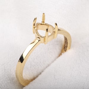 May include: A gold-colored ring setting with a four-prong design to hold a gemstone. The ring has a smooth, polished band and a detailed setting. The ring is shown on a white surface.
