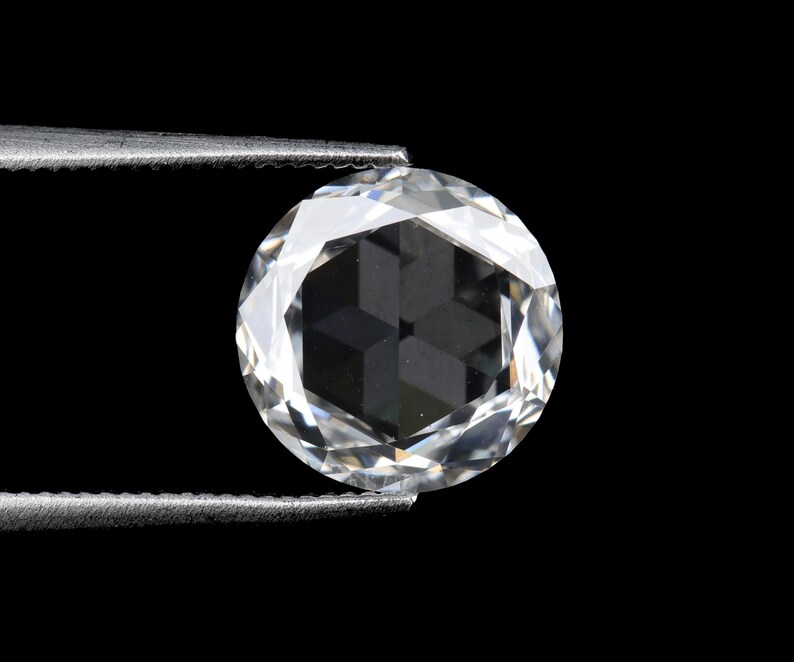 May include: A round, faceted diamond held by tweezers against a black background. The diamond is clear and reflects light, showcasing its many facets and internal structure. The tweezers are silver.