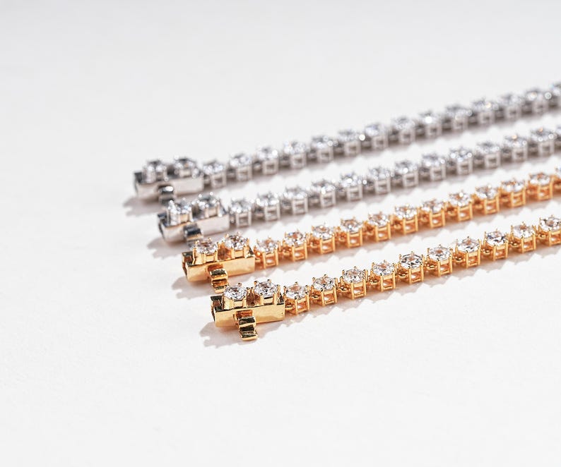 K&ouml;nnte beinhalten: Drei Tennisarmb&auml;nder in Silber- und Goldt&ouml;nen, jedes mit zahlreichen runden, funkelnden Edelsteinen besetzt. Die Armb&auml;nder werden auf einer wei&szlig;en Oberfl&auml;che pr&auml;sentiert, um ihr aufwendiges Design und ihre Brillanz hervorzuheben. Das goldene Armband befindet sich im Vordergrund.
