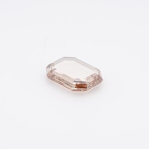 May include: A rectangular, light brown gemstone with an emerald cut. The gem has a clear, transparent appearance with a subtle, warm hue. The facets of the stone reflect light, creating a sparkling effect. The gemstone is set against a white background.