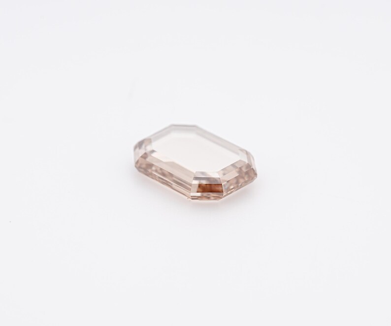 May include: A rectangular, light brown gemstone with an emerald cut. The gem has a clear, transparent appearance with a subtle, warm hue. The facets of the stone reflect light, creating a sparkling effect. The gemstone is set against a white background.