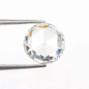 May include: A clear, round-cut diamond held by a pair of silver tweezers. The diamond has multiple facets that reflect light, creating a sparkling effect. The background is plain white, highlighting the gemstone.