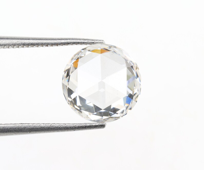 May include: A clear, round-cut diamond held by a pair of silver tweezers. The diamond has multiple facets that reflect light, creating a sparkling effect. The background is plain white, highlighting the gemstone.