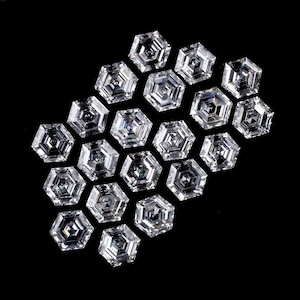 Puede incluir: Una colecci&oacute;n de diecinueve piedras preciosas transparentes e incoloras, con corte hexagonal y dispuestas en grupo. Cada piedra presenta una superficie facetada con un patr&oacute;n geom&eacute;trico que refleja la luz, creando un aspecto brillante y centelleante sobre un fondo negro.