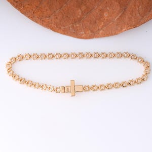 K&ouml;nnte beinhalten: Ein goldfarbenes Armband mit geometrischem Design. Das Armband hat eine Reihe von sechseckigen Formen, die miteinander verbunden sind, mit einem rechteckigen Verschluss. Das Armband wird auf einer wei&szlig;en Oberfl&auml;che pr&auml;sentiert.