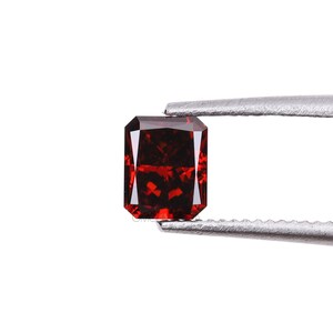 May include: A deep red gemstone with a square shape, held by tweezers. The gemstone is cut into a faceted design.