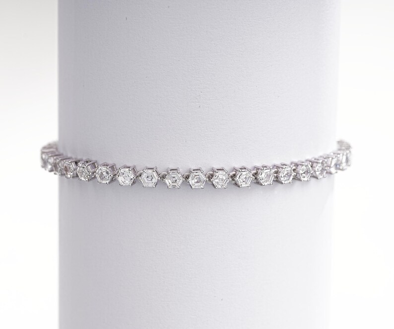 K&ouml;nnte beinhalten: Ein silbernes Armband mit einer Reihe von sechseckigen, klaren Edelsteinen. Das Armband wird vor einem wei&szlig;en Hintergrund pr&auml;sentiert, der sein filigranes Design und seinen Glanz hervorhebt. Die Edelsteine sind gleichm&auml;&szlig;ig angeordnet.