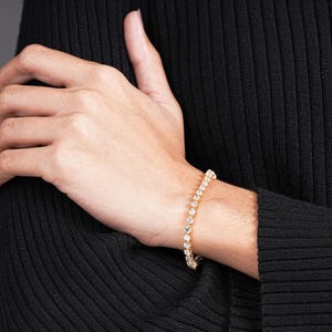K&ouml;nnte beinhalten: Ein goldenes Armband mit zahlreichen klaren, runden Edelsteinen. Das Armband befindet sich am Handgelenk einer Person, die ein schwarzes Langarmshirt tr&auml;gt. Die Edelsteine sind gleichm&auml;&szlig;ig angeordnet.