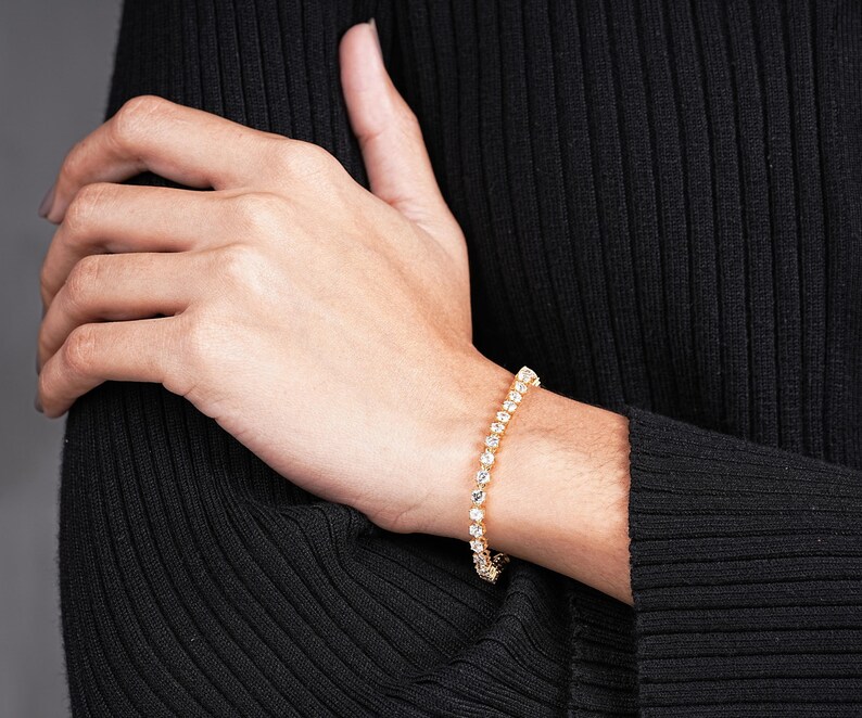 K&ouml;nnte beinhalten: Ein goldenes Armband mit zahlreichen klaren, runden Edelsteinen. Das Armband befindet sich am Handgelenk einer Person, die ein schwarzes Langarmshirt tr&auml;gt. Die Edelsteine sind gleichm&auml;&szlig;ig angeordnet.